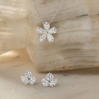 Diamond flower pendant necklace with matching diamond stud earrings displayed on a neutral stone backdrop.