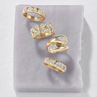 Gold diamond rings with bezel‑set pear and round diamonds arranged on a light purple stone surface.