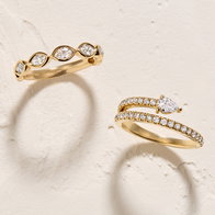 Gold rings featuring marquise and round diamond settings displayed on a textured neutral background.