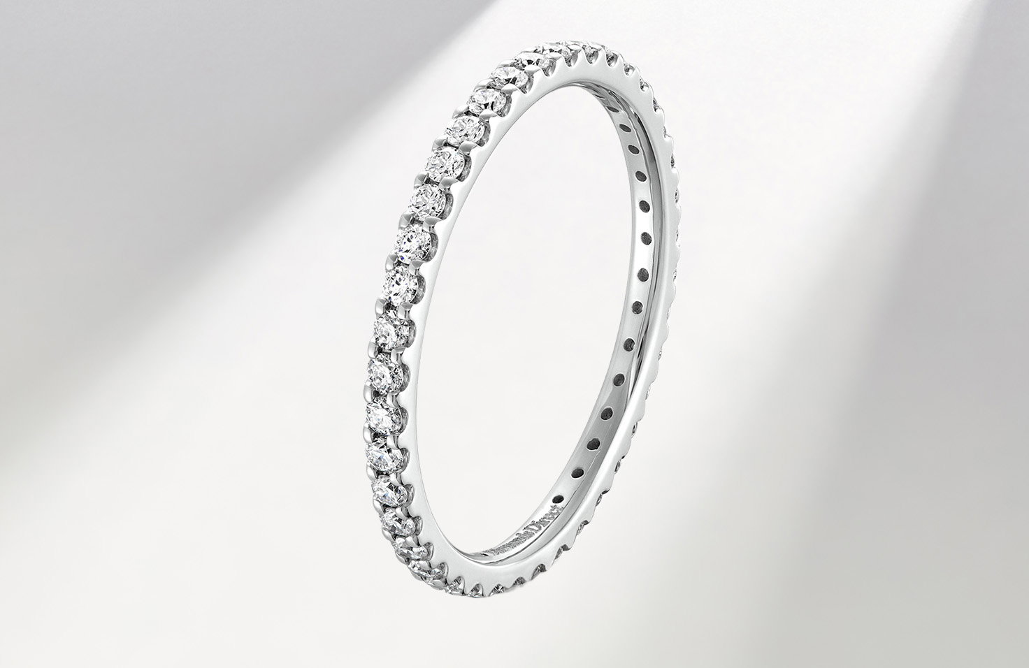 Diamonds Direct Thin white gold diamond eternity ring with round brilliant-cut diamonds encircling the band in a shared-prong setting on a light gray background.