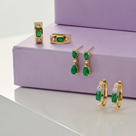 Gold earrings featuring rectangular and teardrop green stone accents displayed on a lavender platform.