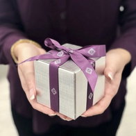 Diamonds Direct silver gift box with purple ribbon featuring the diamond logo, held in both hands.