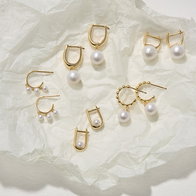 Gold hoop and drop earrings with white pearl accents arranged on a light textured fabric background.