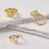 Three yellow gold diamond rings featuring emerald‑cut, pear‑shaped, and pavé‑accented designs displayed on a layered white surface.