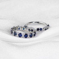 {"en-us":"Two white gold bands with blue sapphires and diamond accents displayed on a white fabric background."}